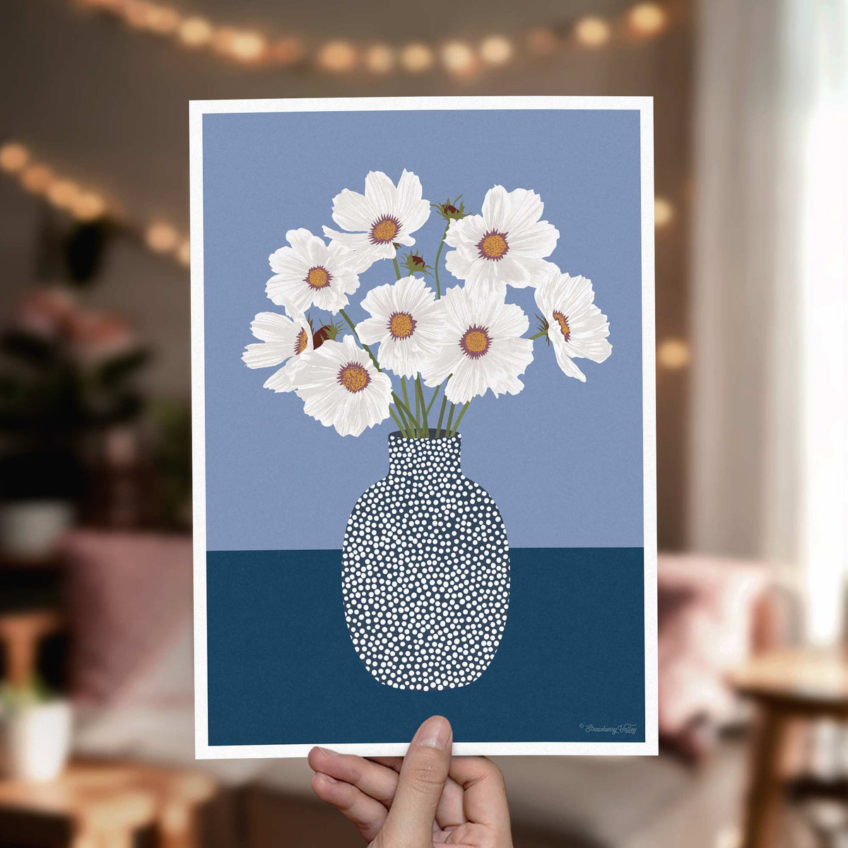 A hand holds a square art print featuring white cosmos flowers in a patterned vase against a split blue background. The print is shown in a softly blurred living room setting with warm string lights in the background.