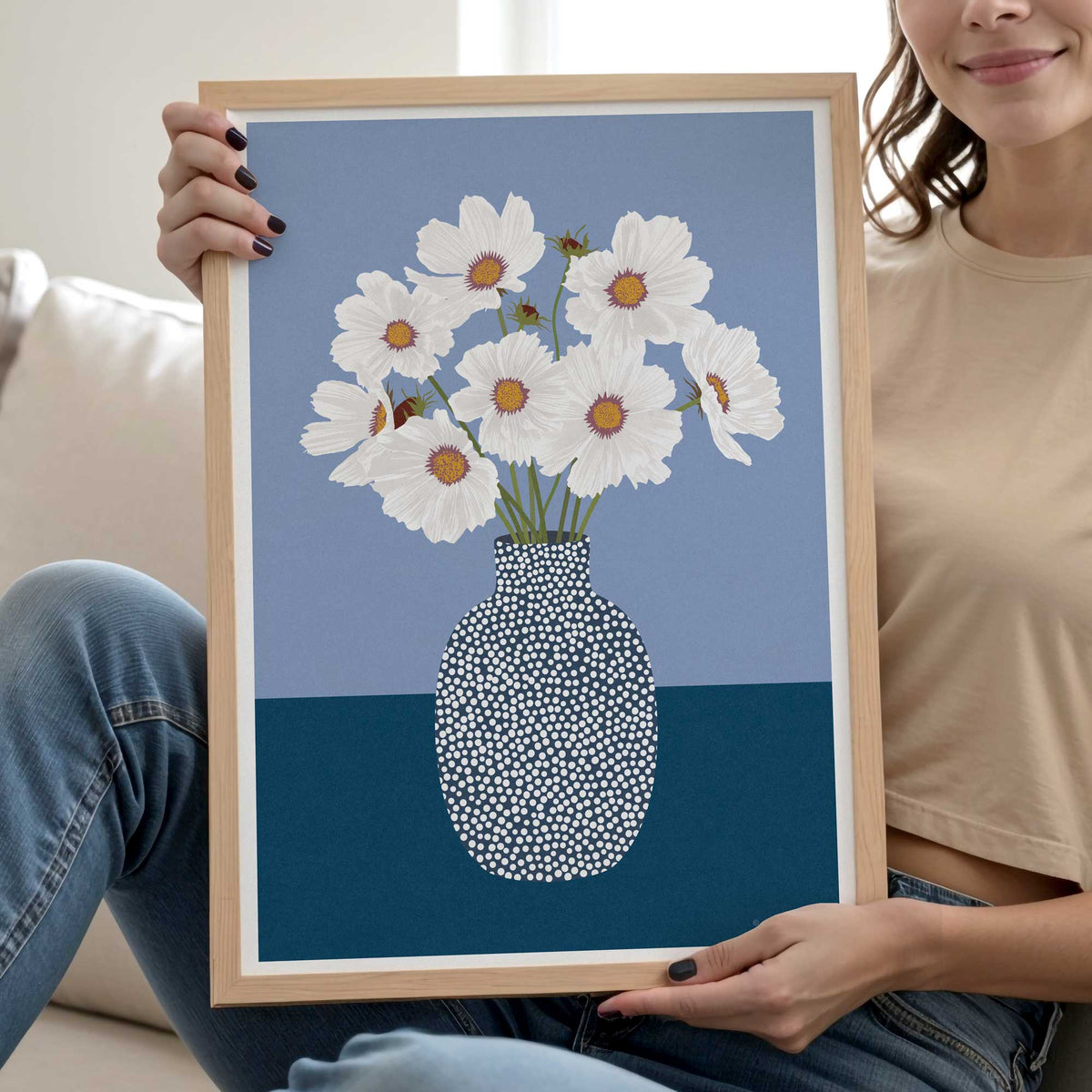 A person holds a light wood framed art print featuring white daisies with orange centers in a speckled blue and navy vase, set against a blue and navy background.