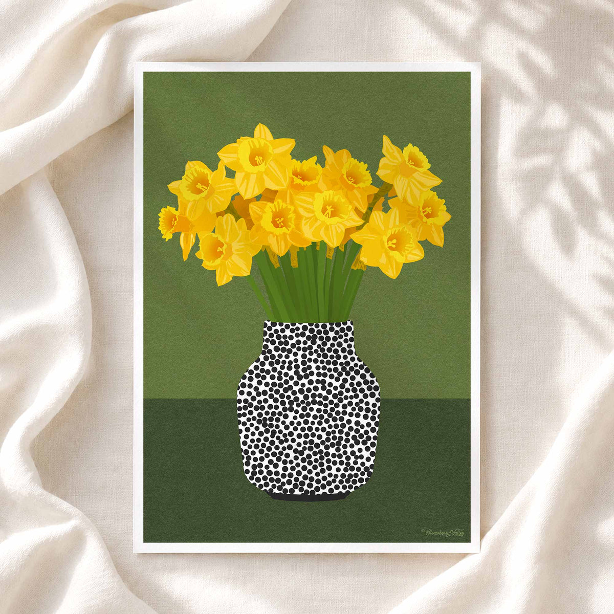 A framed art print featuring yellow daffodils in a black-and-white spotted vase against a muted green background, styled on draped cream fabric.