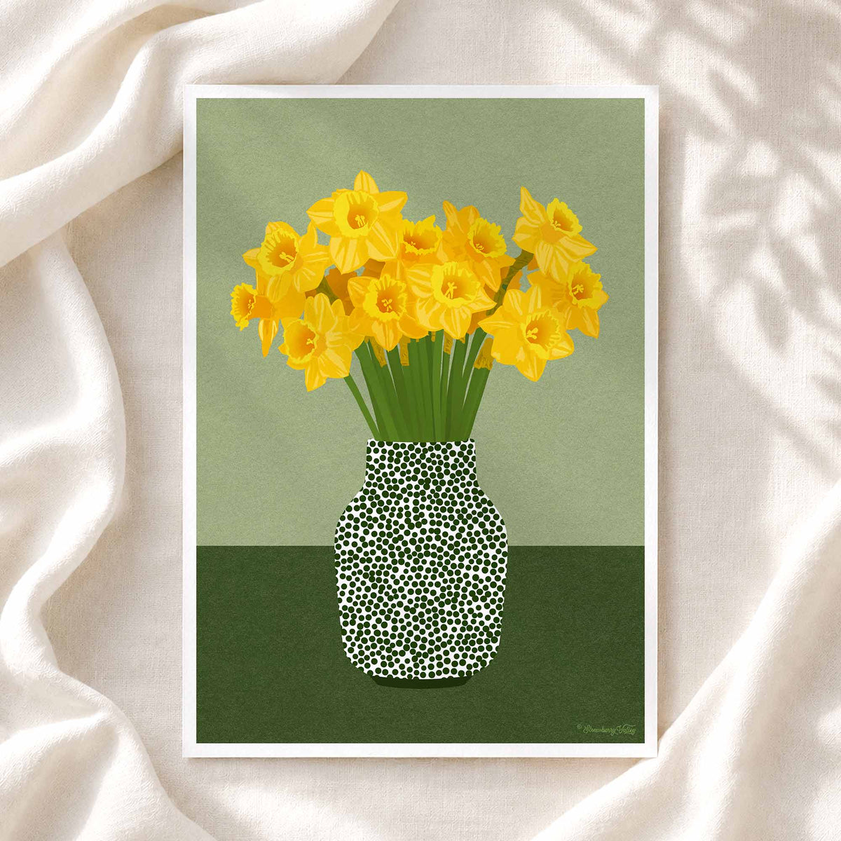 A framed art print featuring yellow daffodils in a black-and-white spotted vase against a muted green background, styled on draped cream fabric.