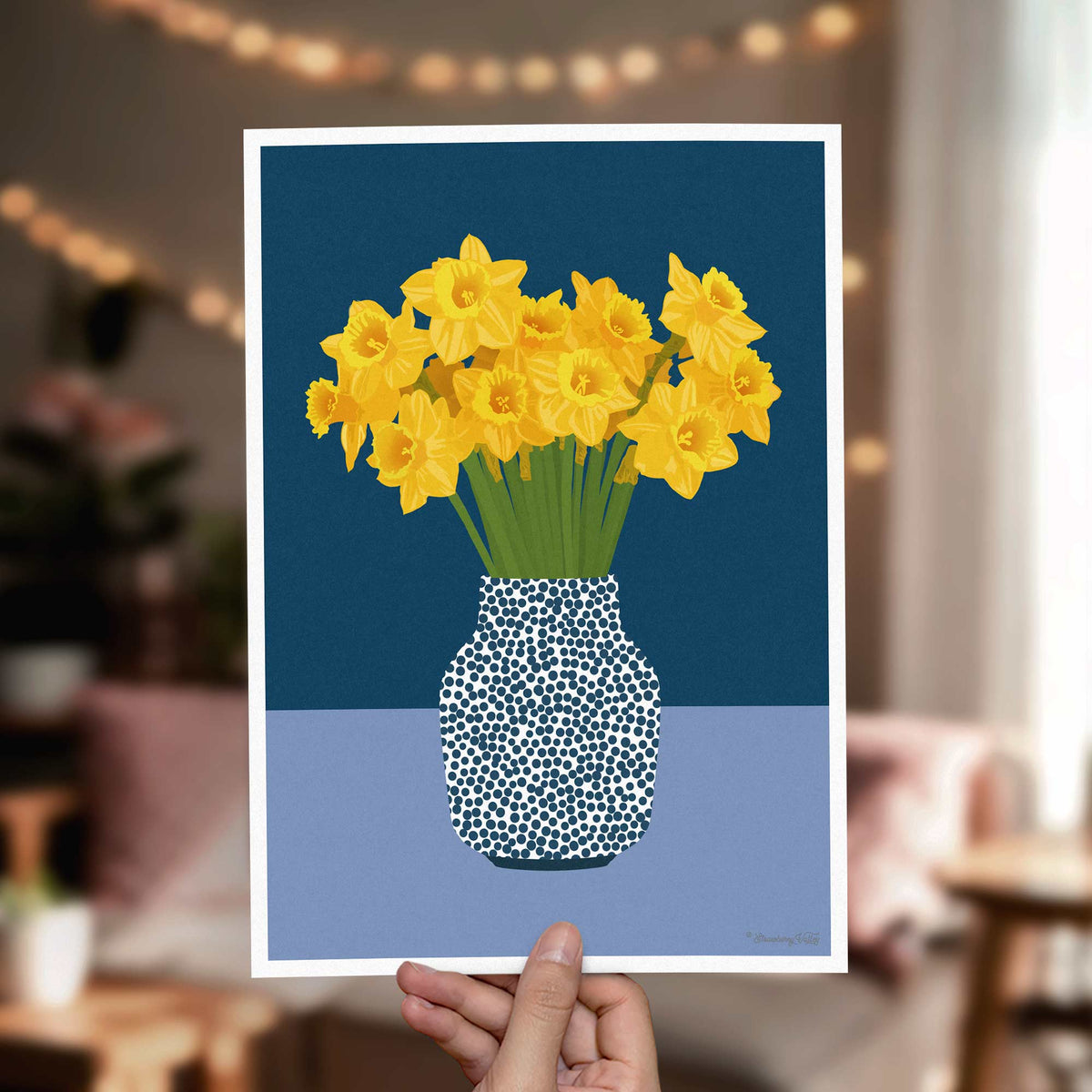 A hand holds a square art print featuring yellow daffodils in a blue-and-white speckled vase against a dark blue and light blue background. The scene is photographed in a softly blurred living room with warm string lights in the background.
