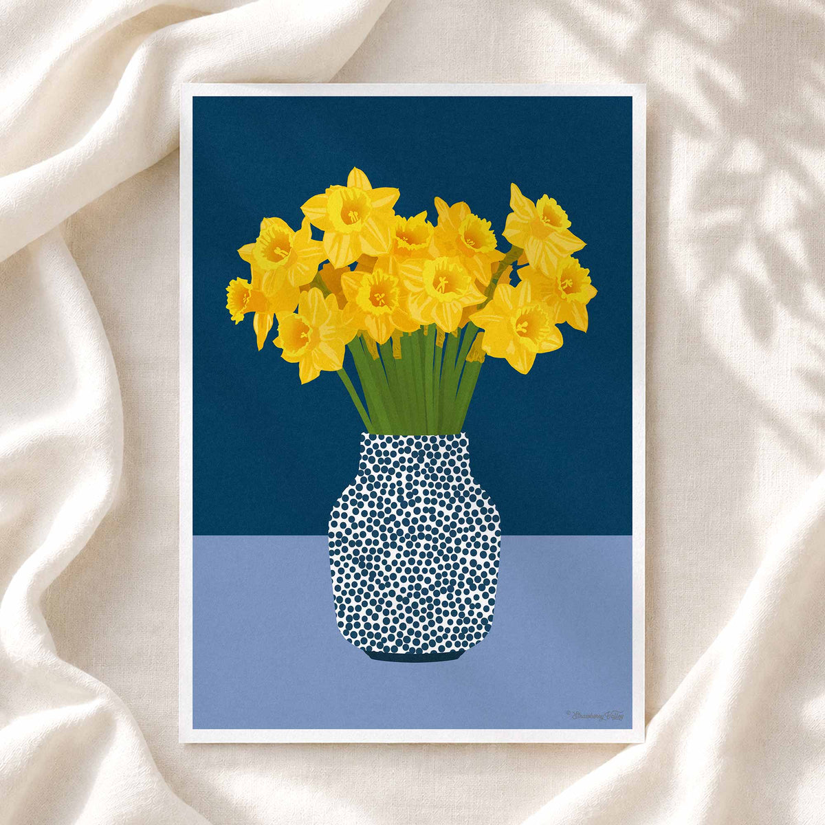 A framed art print featuring a stylized illustration of yellow daffodils in a blue and white speckled vase, displayed on a draped cream fabric background.