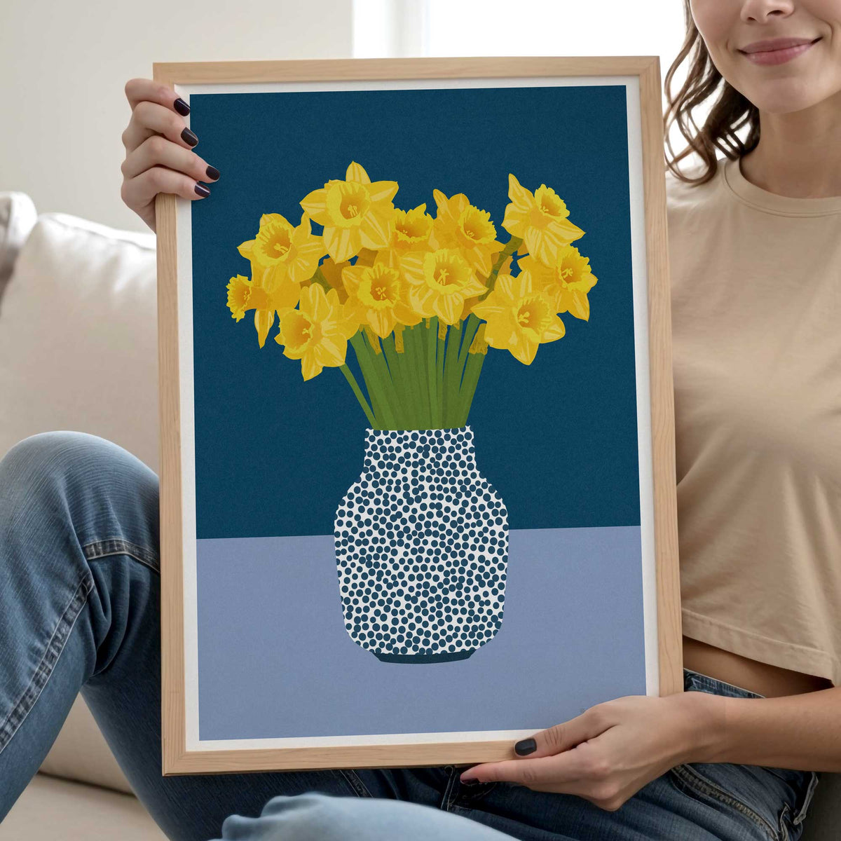 A woman holds a framed art print featuring yellow daffodils in a patterned vase against a dark blue and light blue background. The woman is wearing a beige t-shirt and blue jeans, sitting on a white couch.