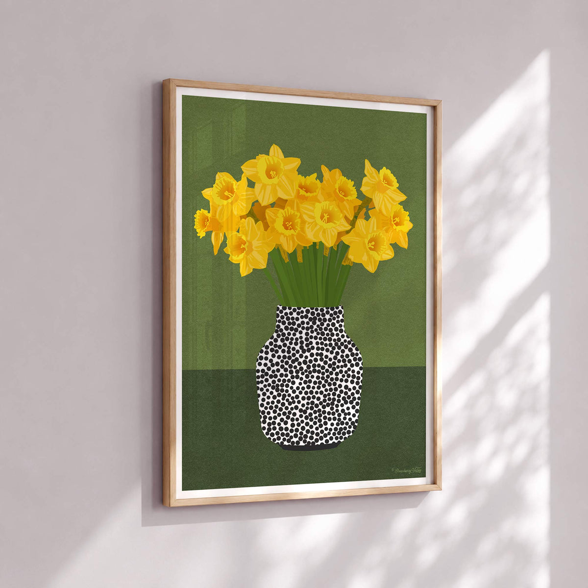 A framed art print featuring yellow daffodils in a black-and-white spotted vase against an olive green background, hung on a light gray wall with soft window shadow patterns.