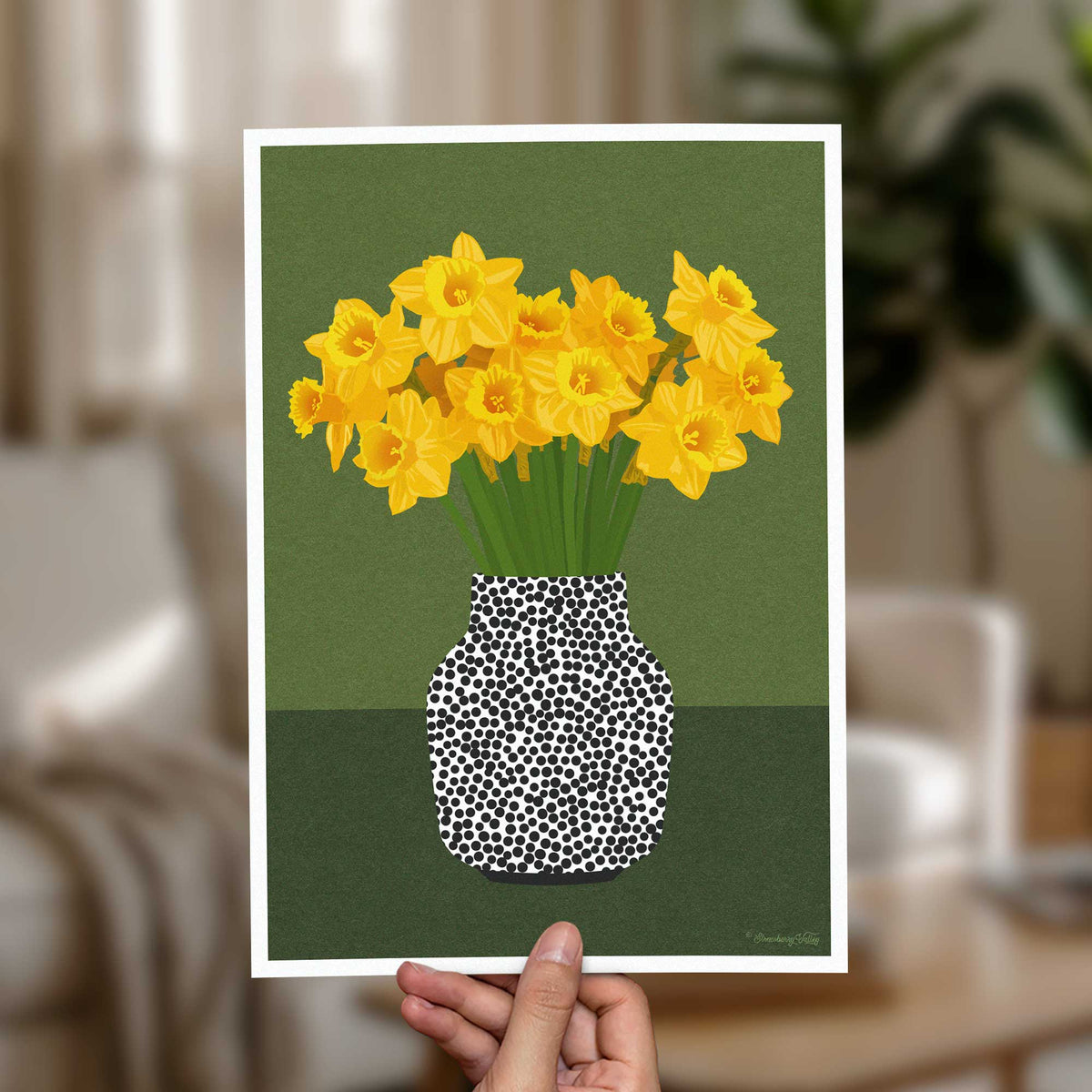 A hand holds a square art print featuring a stylized illustration of yellow daffodils in a black-and-white spotted vase against an olive green background. The room behind is softly blurred, showing a neutral indoor setting.