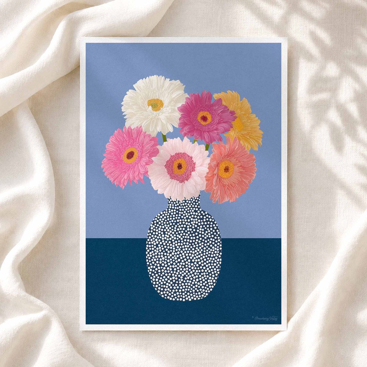A framed art print featuring a colorful bouquet of gerbera daisies in a patterned vase against a blue background, styled on a draped cream fabric surface.