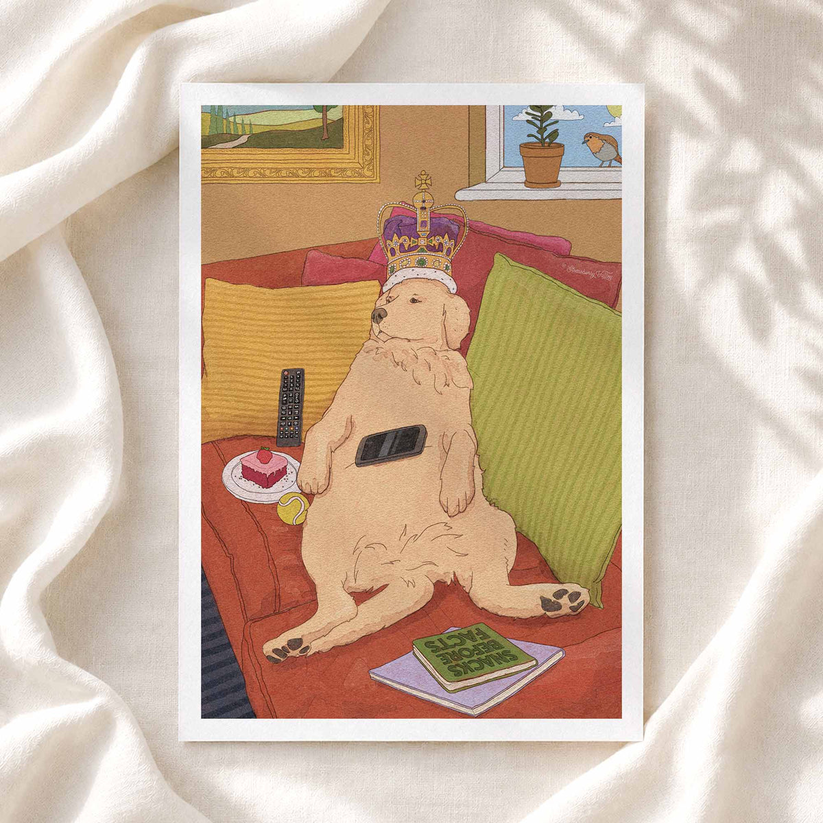 A square art print featuring a humorous illustration of a golden retriever dog wearing a purple and gold crown while lounging on a red couch, surrounded by pillows, a remote control, a phone, a small cake, and a book. The print is displayed flat on draped white fabric with soft shadows.