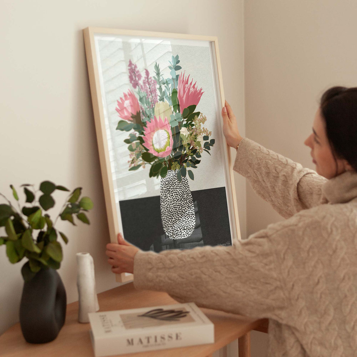 A woman in a beige cable-knit sweater holds a large framed art print featuring a colorful bouquet of pink, purple, and white flowers in a patterned vase. The print is displayed in a light wood frame on a beige wall above a wooden table with a vase of green leaves and a book.