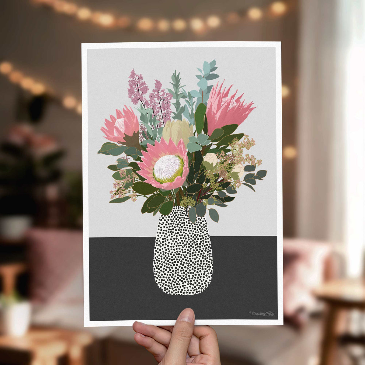 A hand holds a vertical art print featuring a stylized floral bouquet in a patterned vase, set against a softly blurred indoor background with warm string lights.