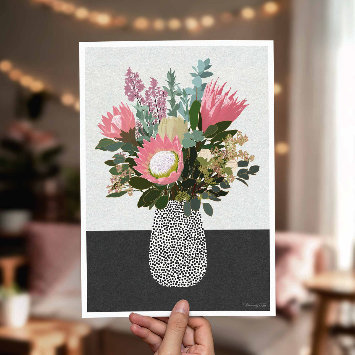 A hand holds a square art print featuring a stylized floral bouquet in a speckled vase, with a softly blurred indoor room in the background.