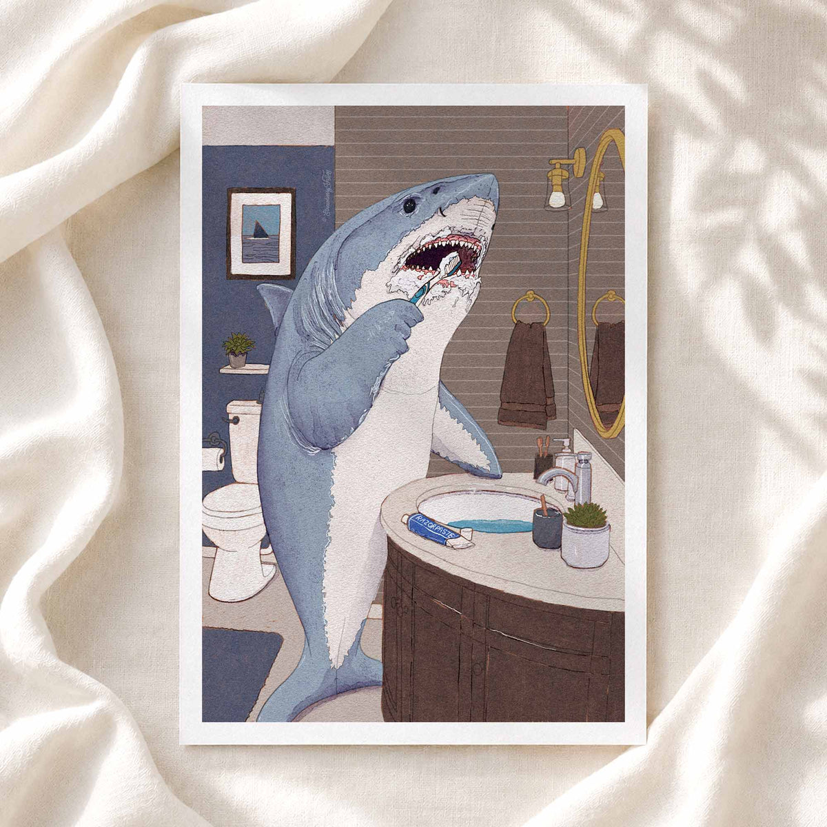 A framed art print featuring a humorous illustration of a great white shark brushing its teeth in a bathroom, styled on a draped cream-colored fabric background.