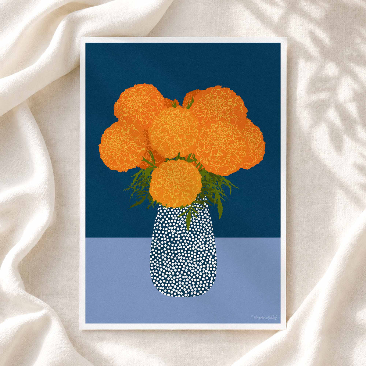 A framed art print featuring a stylized illustration of orange marigold flowers in a patterned blue and white vase, set against a dark blue and light blue background, displayed on draped white fabric.