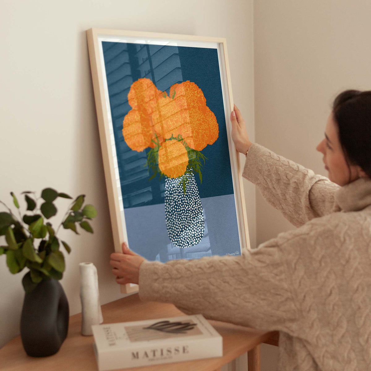 A woman in a beige cable-knit sweater holds a large framed art print against a neutral beige wall. The print features a stylized, pixelated orange flower bouquet in a patterned blue and white vase.