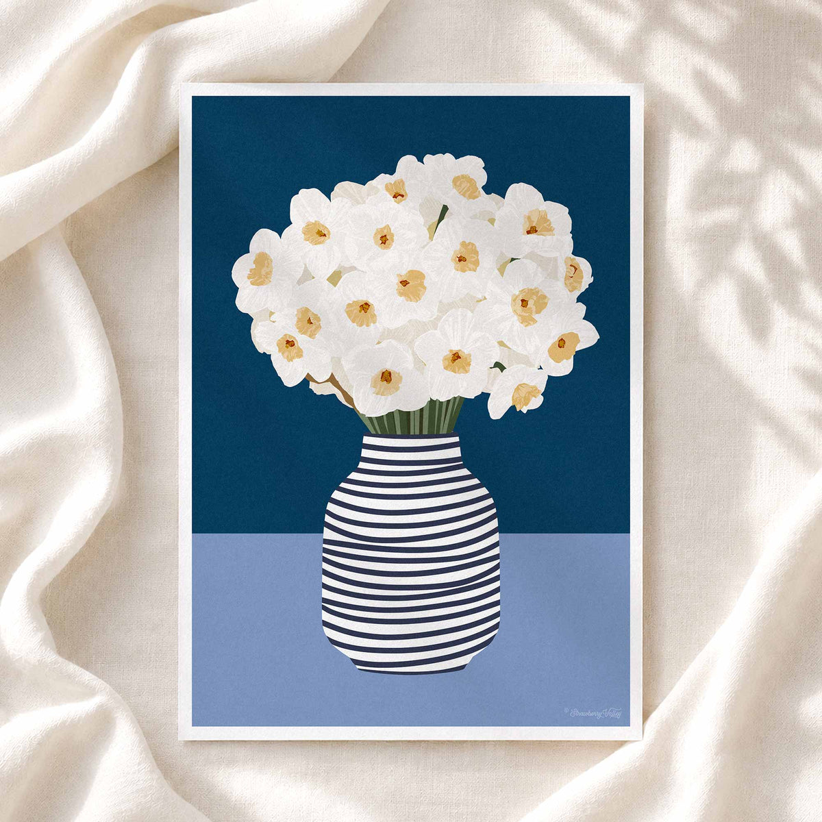 A framed art print featuring white daffodils in a navy and white striped vase, set against a dark blue and light blue background, styled on a draped cream fabric backdrop.
