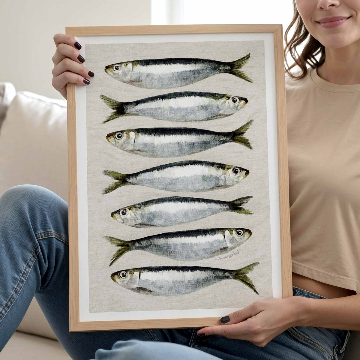 A framed art print featuring seven watercolor illustrations of silver and black fish on a textured off-white background, held by a person sitting on a couch.