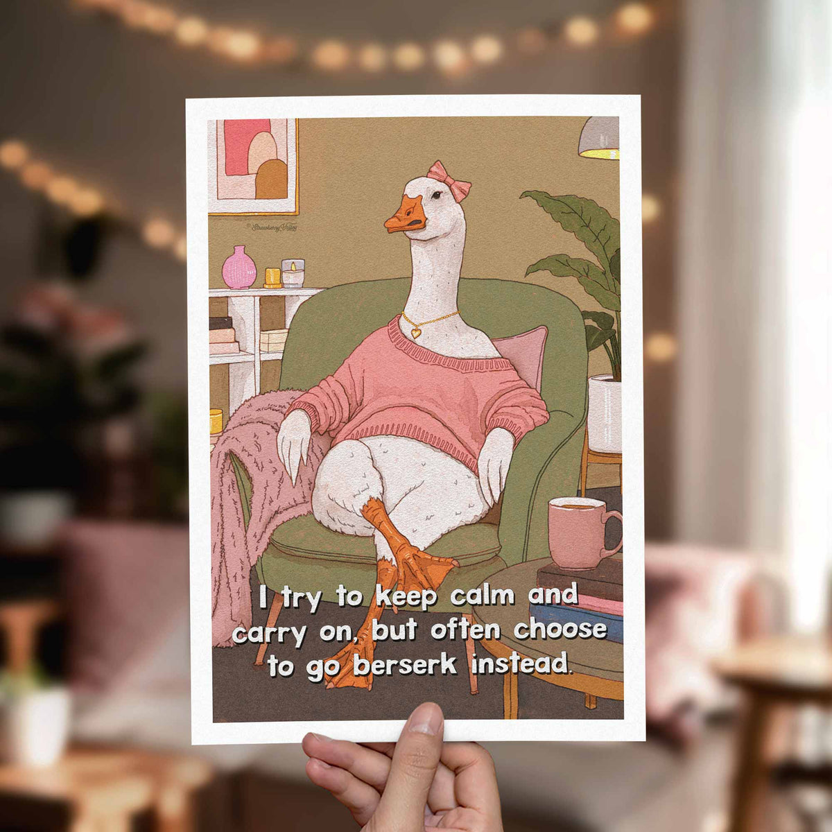 A hand holds a white art print featuring an illustrated goose sitting in a green armchair, wearing a pink sweater and white socks, with a mug and books on a side table. The print includes the text, 'I try to keep calm and carry on, but often choose to go berserk instead.