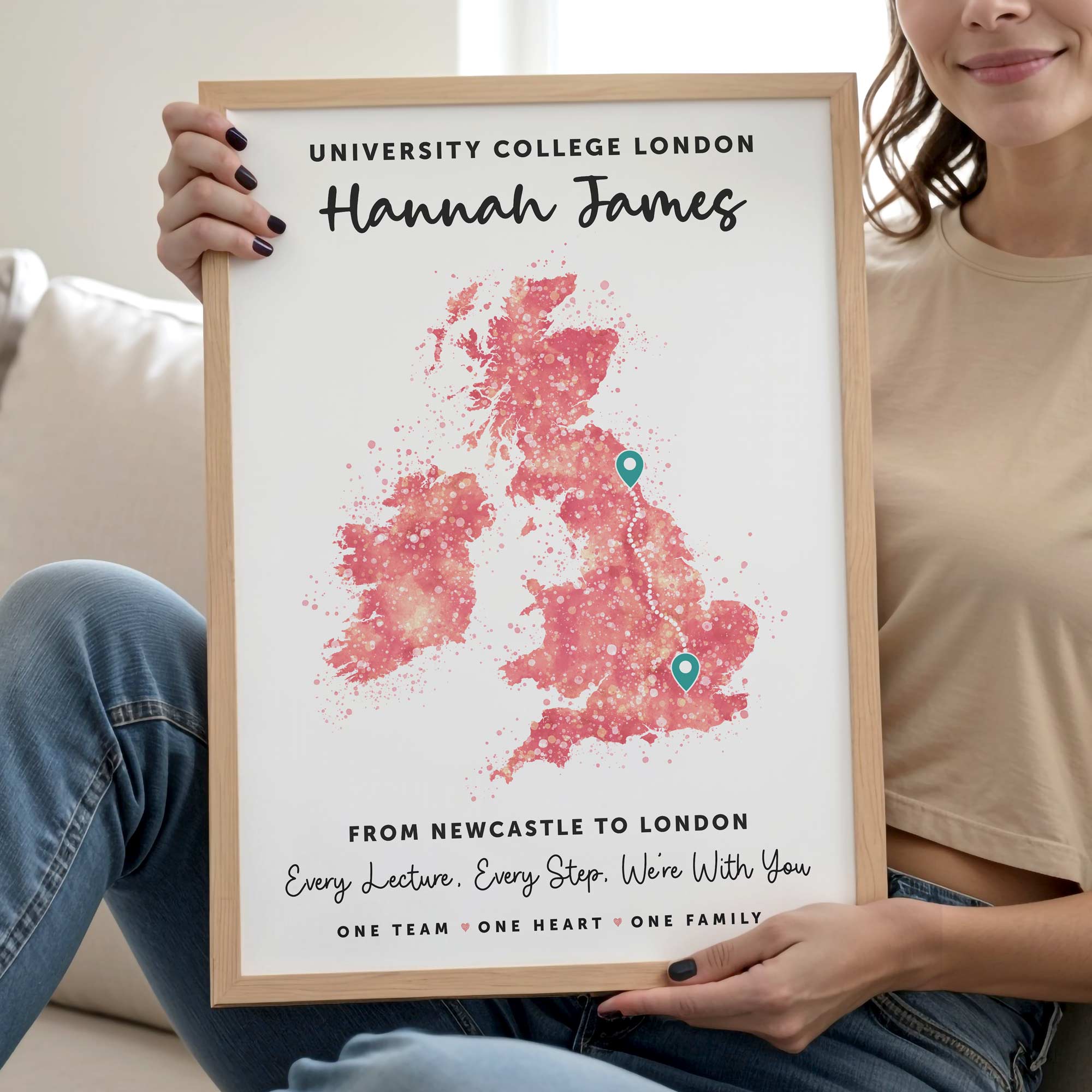 A woman in a beige t-shirt and blue jeans holds up a personalized wooden-framed print. The print features a watercolor-style pink map of the United Kingdom with two teal pin markers, custom text reading 'Hannah James' and 'University College London 2026', and a supportive message at the bottom.