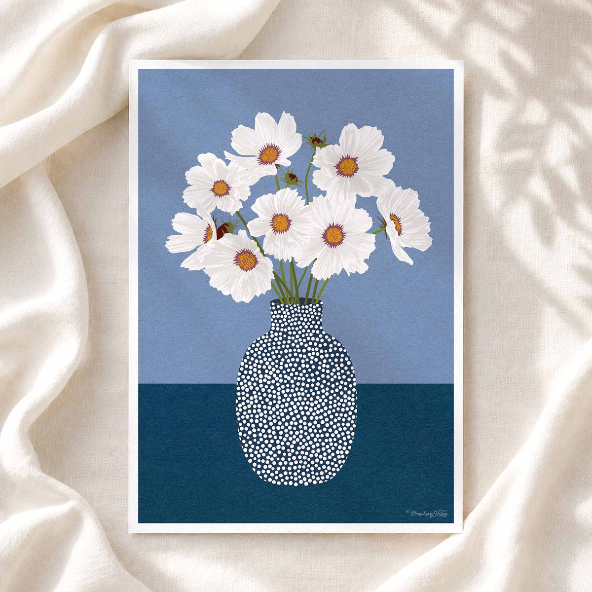 A framed art print featuring a stylized illustration of white cosmos flowers with yellow-orange centers in a patterned blue and navy vase, displayed on draped cream fabric.