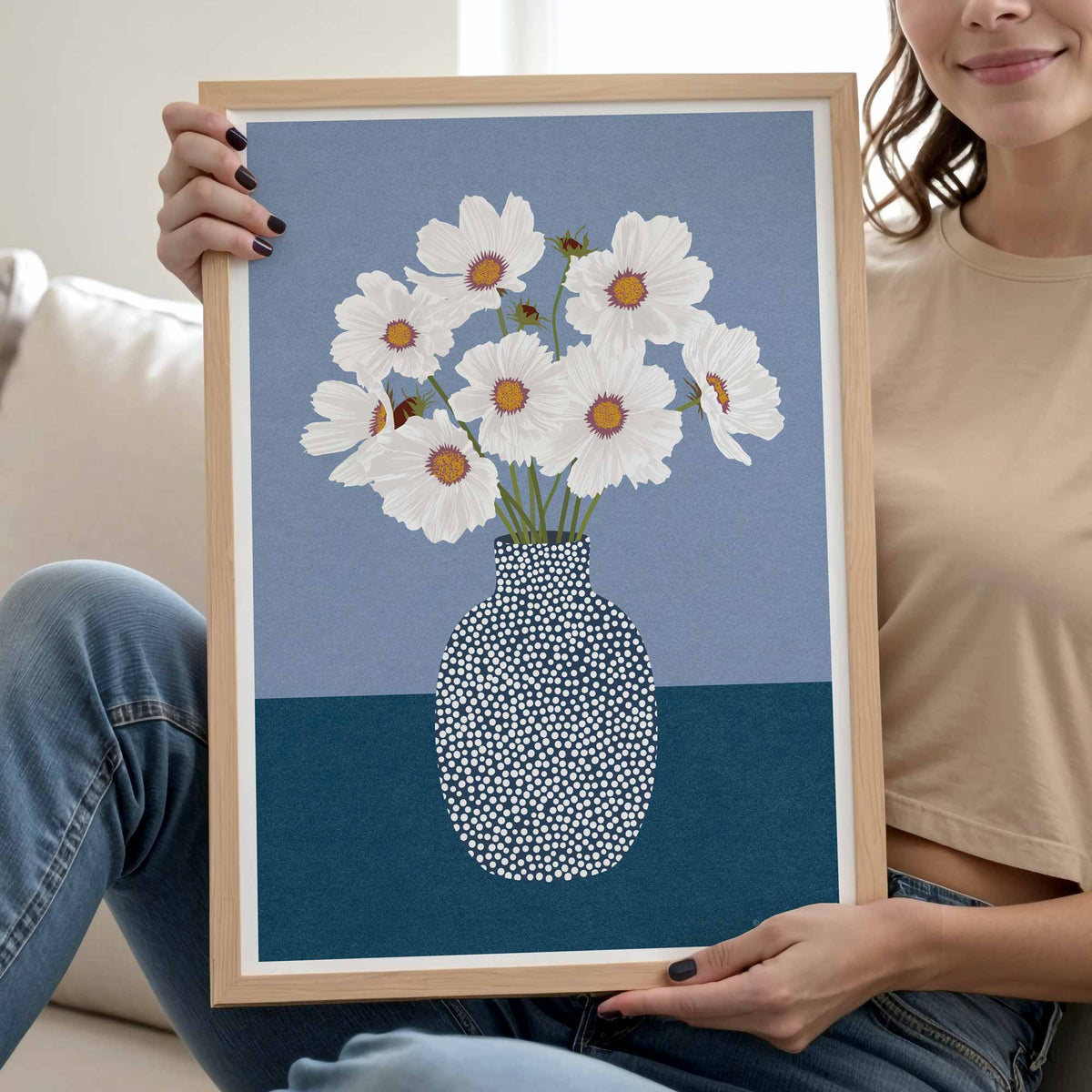A person holds a framed art print featuring white daisies with orange centers in a patterned white and blue vase against a blue background. The print is housed in a light wood frame, and the person is seated on a light-colored sofa.