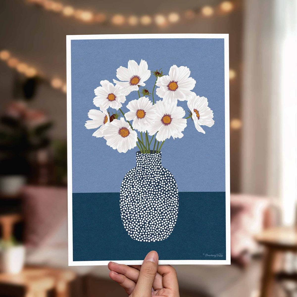 A hand holds a square art print featuring white daisies with orange centers in a speckled blue and navy vase against a blue background. The print is shown in a softly blurred living room setting with warm string lights and furniture in the background.