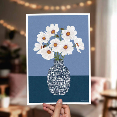 A hand holds a square art print featuring white daisies with orange centers in a speckled blue and navy vase against a blue background. The print is shown in a softly blurred living room setting with warm string lights and furniture in the background.