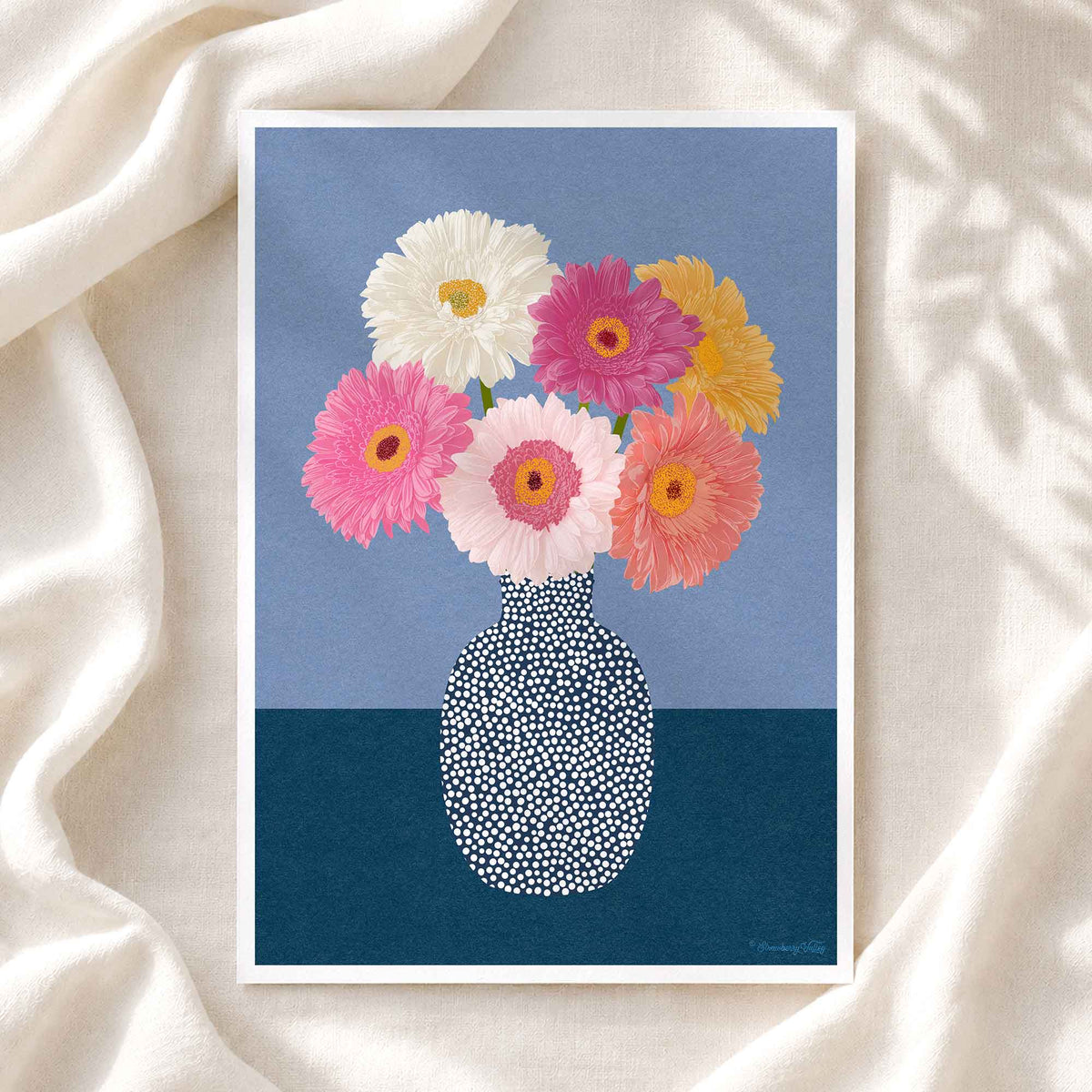 A framed art print featuring a colorful bouquet of gerbera daisies in a patterned vase, displayed on a draped cream fabric background.