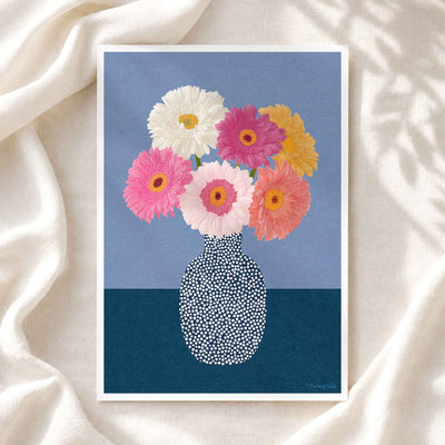 A framed art print featuring a colorful bouquet of gerbera daisies in a patterned vase, displayed on a draped cream fabric background.