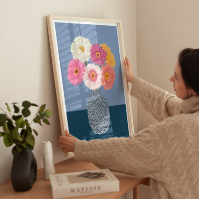 A woman in a beige cable-knit sweater holds a large framed art print featuring a colorful bouquet of flowers in a patterned vase against a blue background, leaning it on a wooden table in a styled room.
