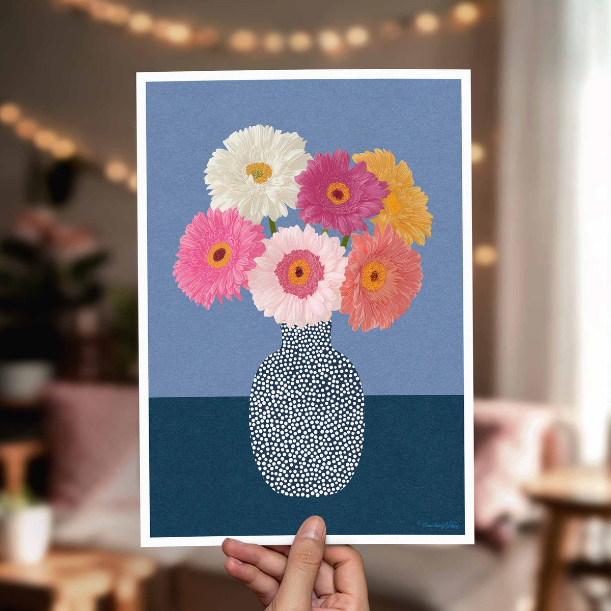 A hand holds a square art print featuring a bouquet of colorful flowers in a patterned vase against a blue background. The blurred room behind suggests a cozy indoor setting with warm lights.