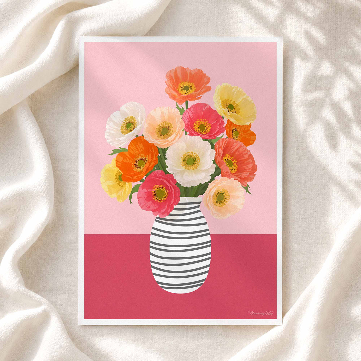 A framed art print featuring a colorful bouquet of flowers in a striped vase, displayed on draped cream fabric with soft leaf shadows.