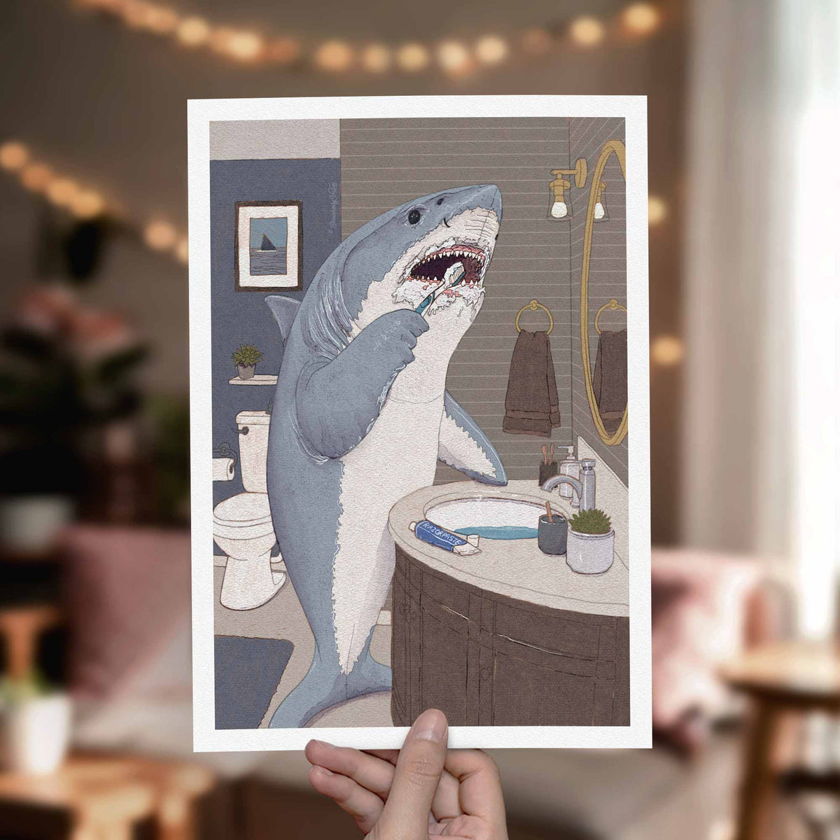 A hand holds a white-bordered art print featuring a humorous illustration of a gray and white shark brushing its teeth in a bathroom. The background is a softly blurred real-world bathroom setting with warm string lights.