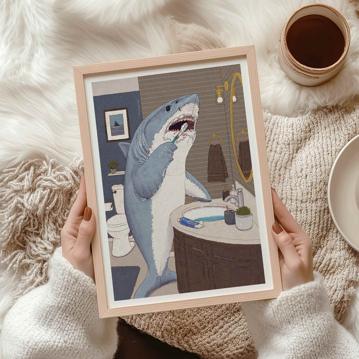 A person holds a light wood framed art print featuring a humorous illustration of a great white shark brushing its teeth in a bathroom. The scene is styled on a cozy beige knit blanket over a white faux fur rug, with a cup of coffee and a plate nearby.