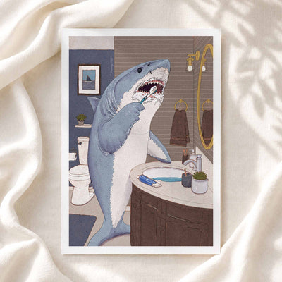 A framed art print featuring a humorous illustration of a great white shark brushing its teeth in a bathroom, styled on a draped cream-colored fabric background.