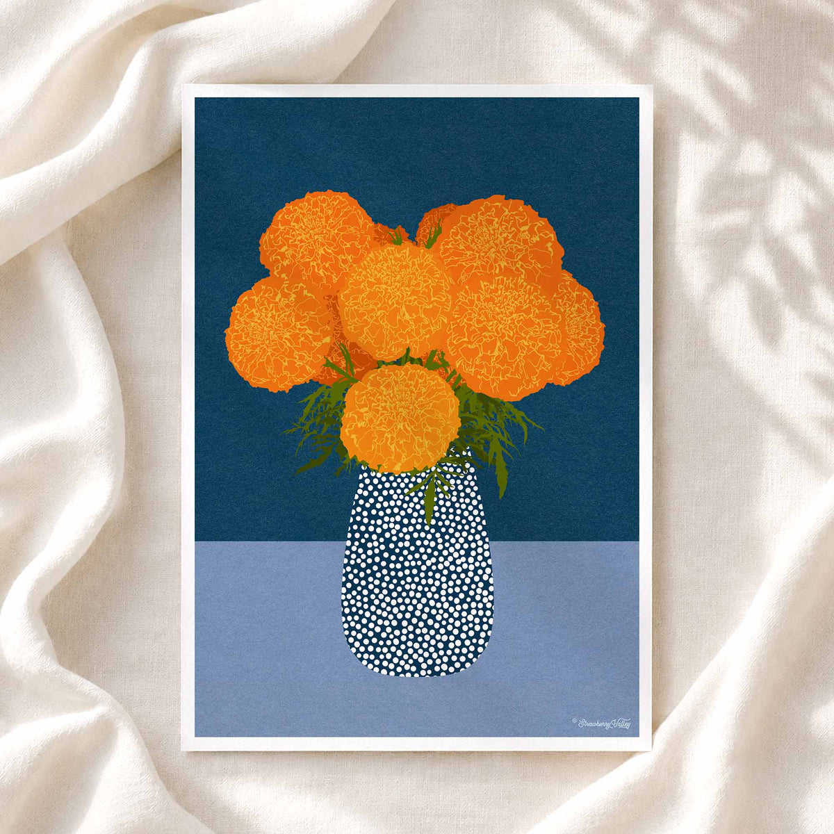 A framed art print featuring a stylized illustration of orange marigold flowers in a patterned blue and white vase, set against a dark blue and light blue background, displayed on draped white fabric.