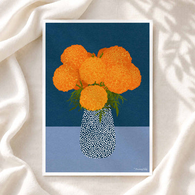 A framed art print featuring a stylized illustration of orange marigold flowers in a patterned blue and white vase, set against a dark blue and light blue background, displayed on draped white fabric.