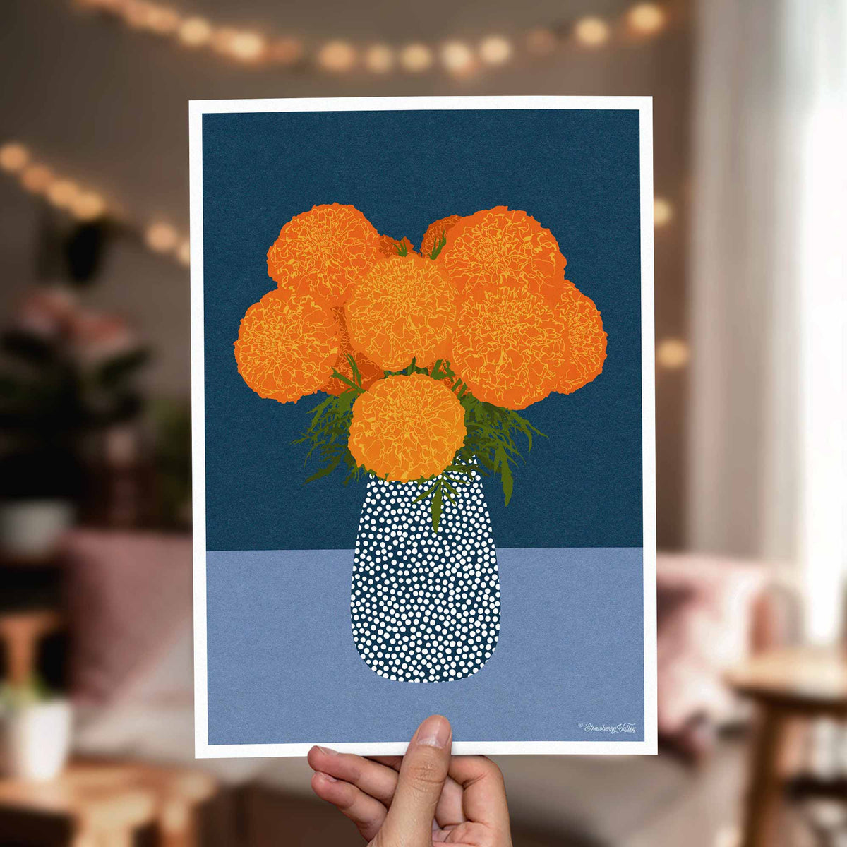 A hand holds a square art print featuring a stylized orange flower bouquet in a white dotted vase against a dark blue and light blue background. The scene is photographed in a softly blurred living room with warm string lights and furniture in the background.