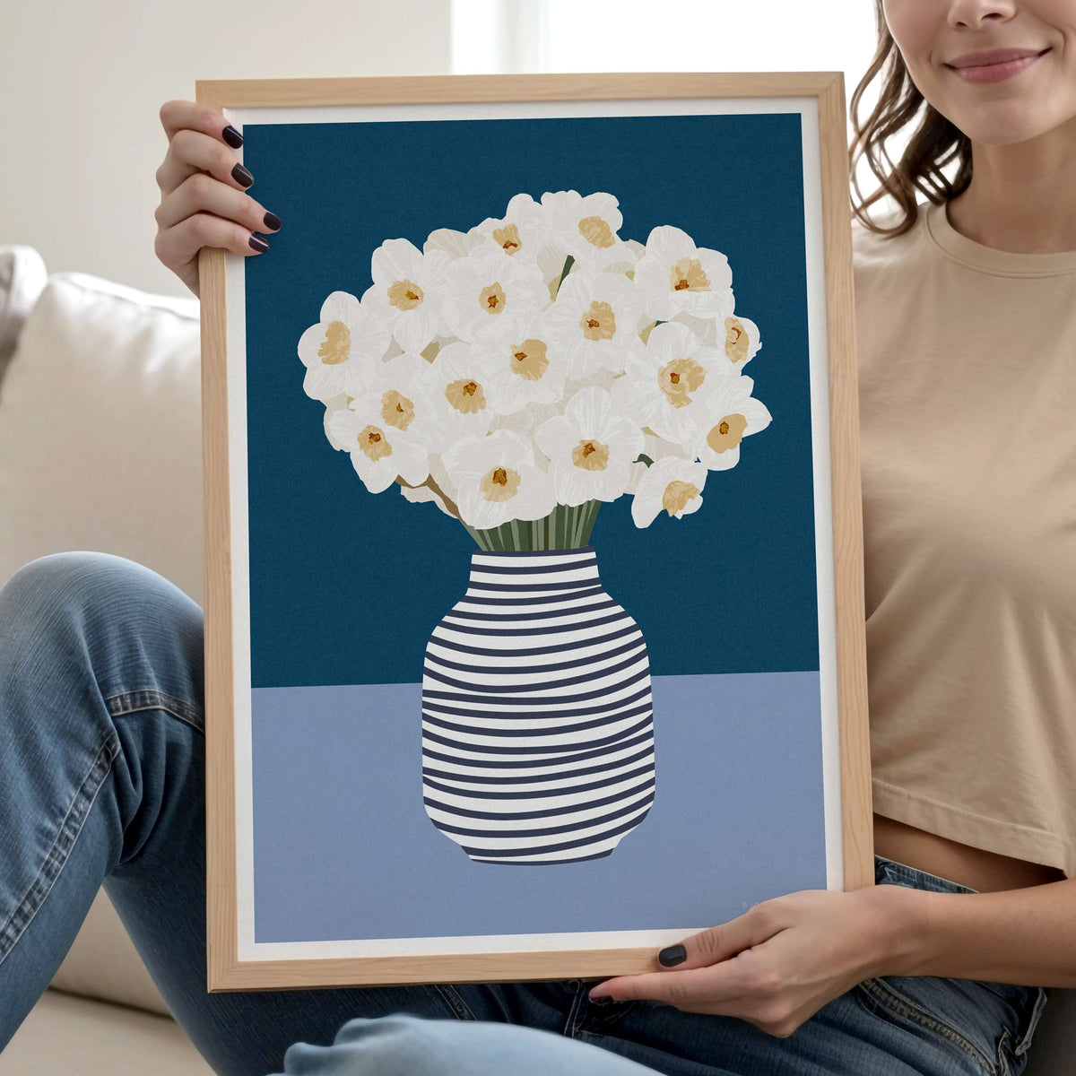 A woman holds a framed art print featuring white daffodils in a black-and-white striped vase against a dark blue and light blue background. The print is housed in a light natural wood frame, and the woman is seated on a sofa wearing a beige crop top and blue jeans.