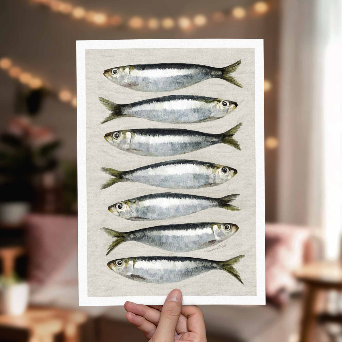 A hand holds a white art print featuring seven horizontal illustrations of silver and black fish on a light background, with a softly blurred indoor room in the background.