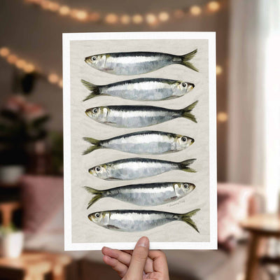 A hand holds a white art print featuring seven horizontal illustrations of silver and black fish on a light background, with a softly blurred indoor room in the background.