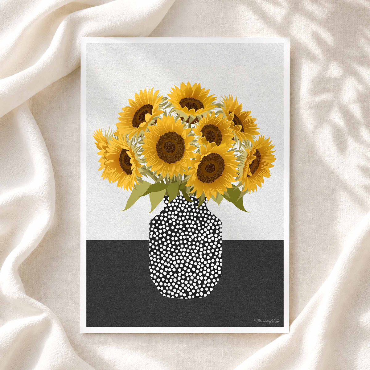 A framed art print featuring a bouquet of yellow sunflowers in a black-and-white dotted vase, styled on draped cream fabric with soft leaf shadows.