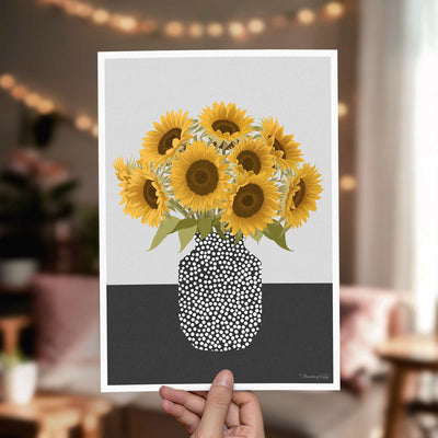 A hand holds a vertical art print featuring a stylized illustration of yellow sunflowers in a black-and-white dotted mason jar vase against a light gray background. The print is shown in a softly blurred living room setting with warm string lights and furniture in the background.