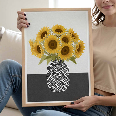 A woman holds a light wood framed art print featuring a bouquet of yellow sunflowers in a white and dark gray vase with a black-and-white dotted pattern. She is sitting on a white couch wearing a beige crop top and blue jeans.
