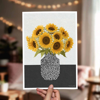 A hand holds a vertical art print featuring a stylized bouquet of yellow sunflowers in a black-and-white dotted vase against a split light gray and dark gray background. The scene is photographed in a softly blurred living room with warm string lights in the background.