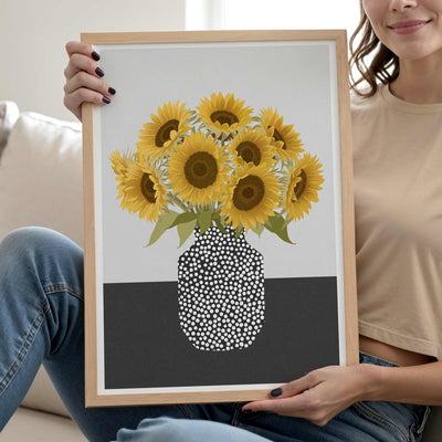 A woman holds a light wood framed art print featuring a bouquet of yellow sunflowers in a black and white dotted vase, set against a neutral indoor background.