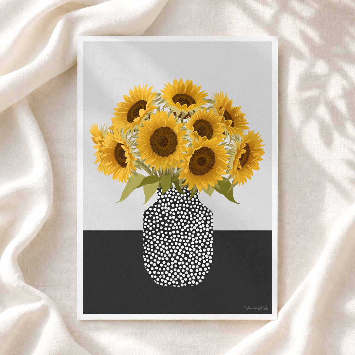 A framed art print featuring a bouquet of yellow sunflowers in a black-and-white dotted vase, displayed on draped cream fabric with soft leaf shadows.