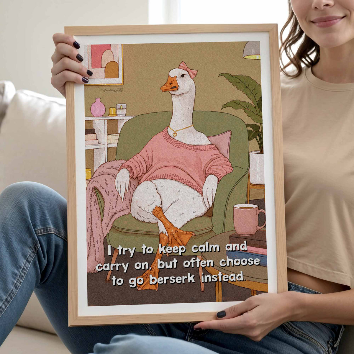 A person holds up a framed art print featuring a white goose wearing a pink sweater and sitting in an armchair with the text, 'I try to keep calm and carry on, but often choose to go berserk instead.' The background shows a cozy living room setting with a sofa and plant.