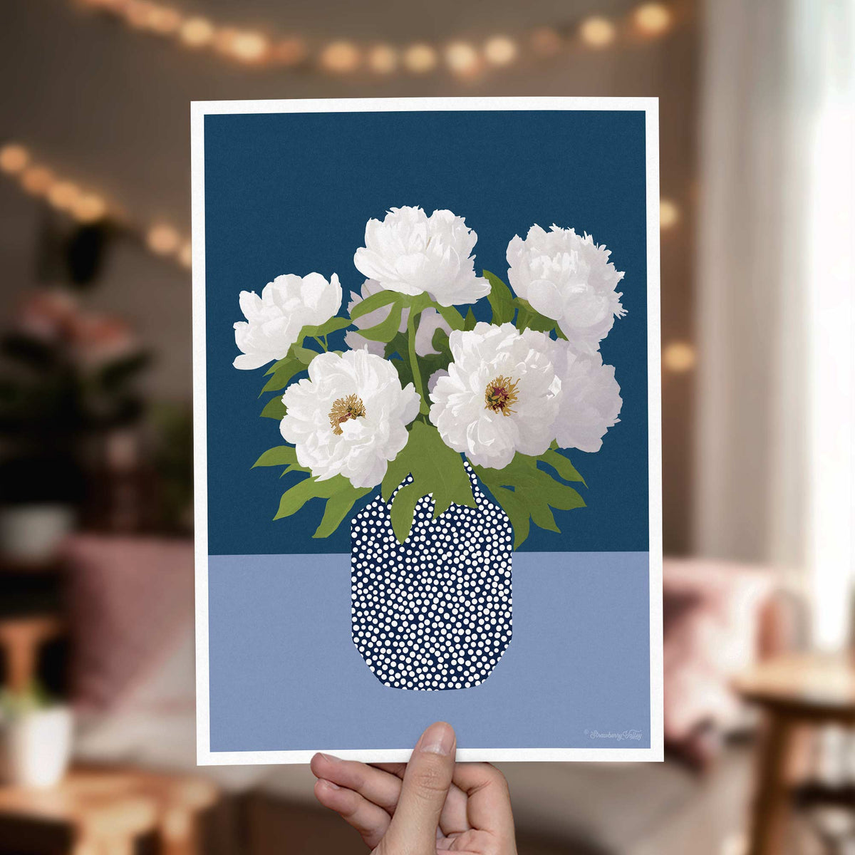 A hand holds a vertical art print featuring white peonies in a patterned vase against a dark blue and light blue background. The scene is set in a softly blurred indoor environment with warm string lights in the background.