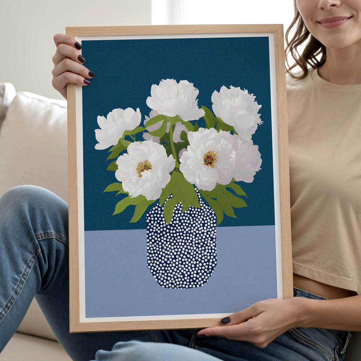 A woman holds a light wood framed art print featuring white peonies in a black-and-white dotted vase against a dark teal and light blue background.