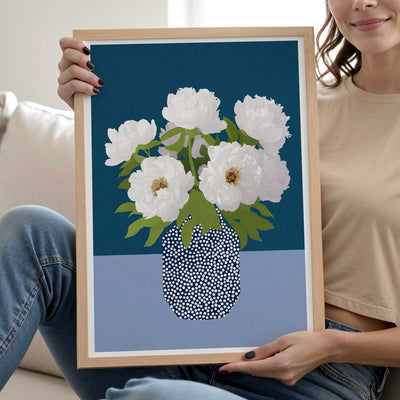 A woman holds a light wood framed art print featuring white peonies in a black-and-white dotted vase against a dark teal and light blue background.