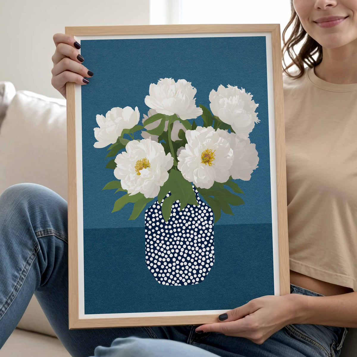 A person sitting on a couch holds up a framed art print featuring white peony flowers in a blue and white dotted vase against a dark blue background.